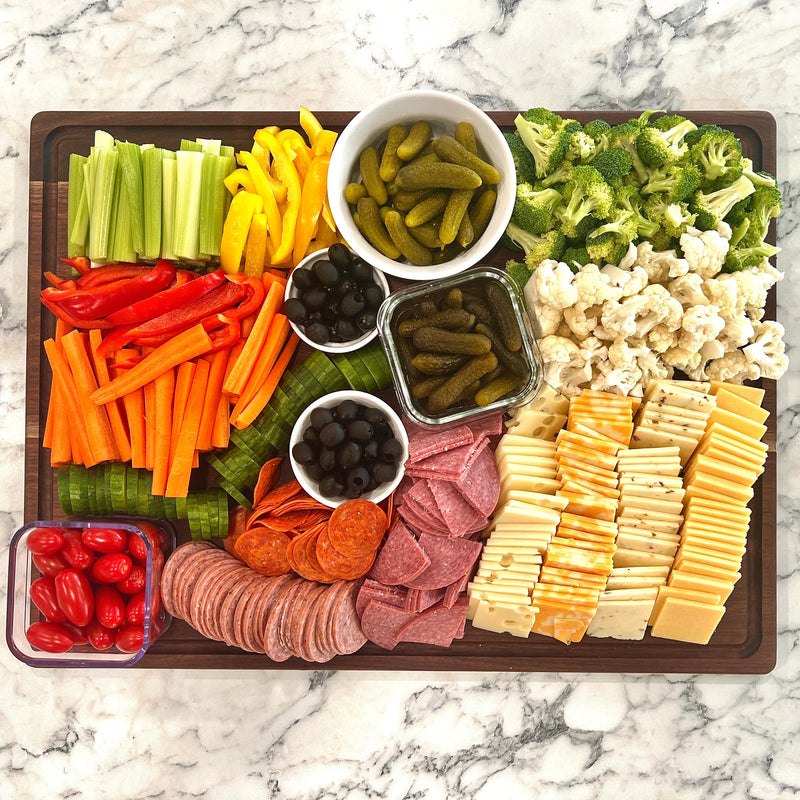 Overhead view of a walnut serving board with sliced meats, cheeses, vegetables, pickles and olives