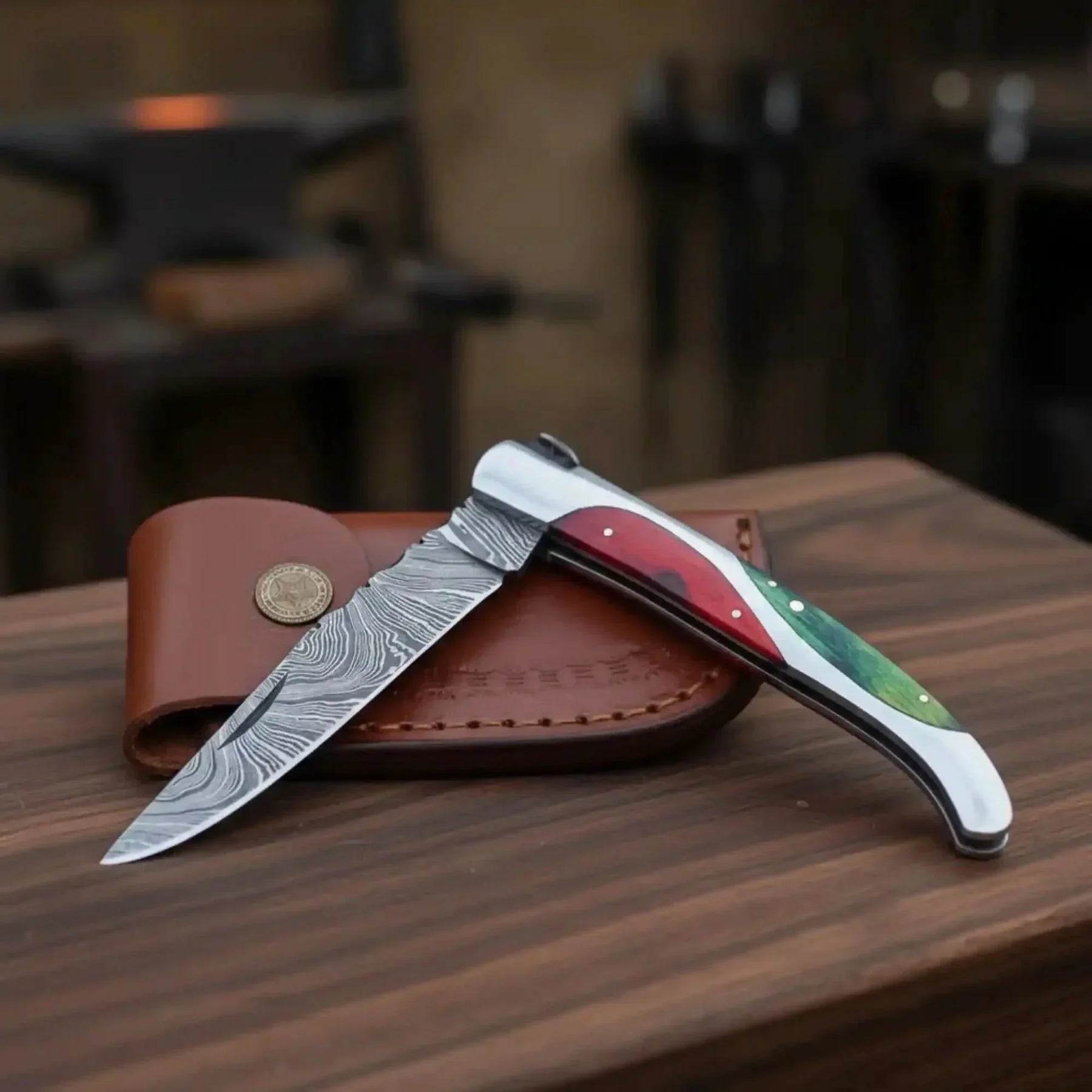 Open folding pocket knife with a patterned blade, red-and-green handle and brown leather pouch