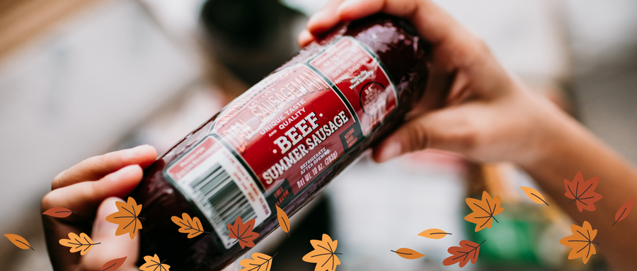 Hands holding wrapped beef summer sausage above a surface decorated with autumn leaves