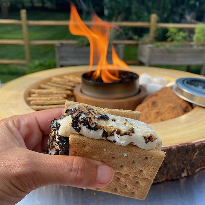 Person holding a prepared s’more beside an outdoor bonfire