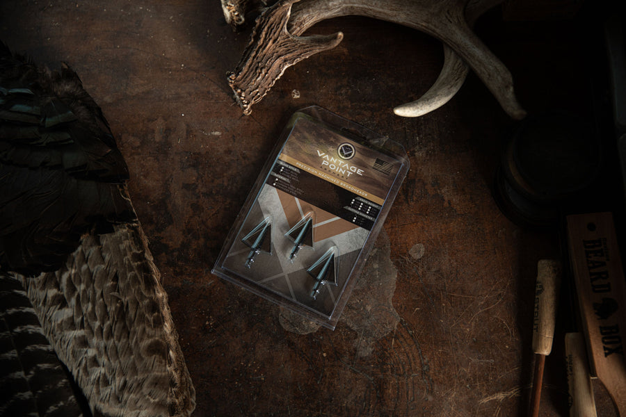 Packaged double-bevel broadheads beside hunting equipment