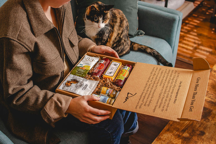 Person opening a Dan The Sausageman gift box with a cat sitting nearby