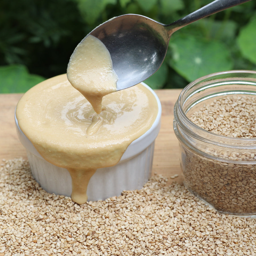 Tahini dripping from a spoon into a bowl beside a glass jar of sesame seeds
